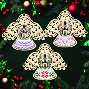 Peut inclure: Trois ornements en bois en forme d'ange avec des motifs de point de croix. Chaque ange pr&eacute;sente une palette de couleurs diff&eacute;rente : violet, rose et vert. Les anges sont entour&eacute;s de d&eacute;corations de No&euml;l, dont du houx, du pin et des boules.