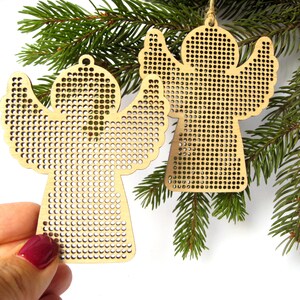 May include: Two wooden angel ornaments with perforated designs, hanging from twine, displayed on a green pine branch. The angels have outstretched wings and are made of light-colored wood. One is held by a hand with a maroon nail polish.