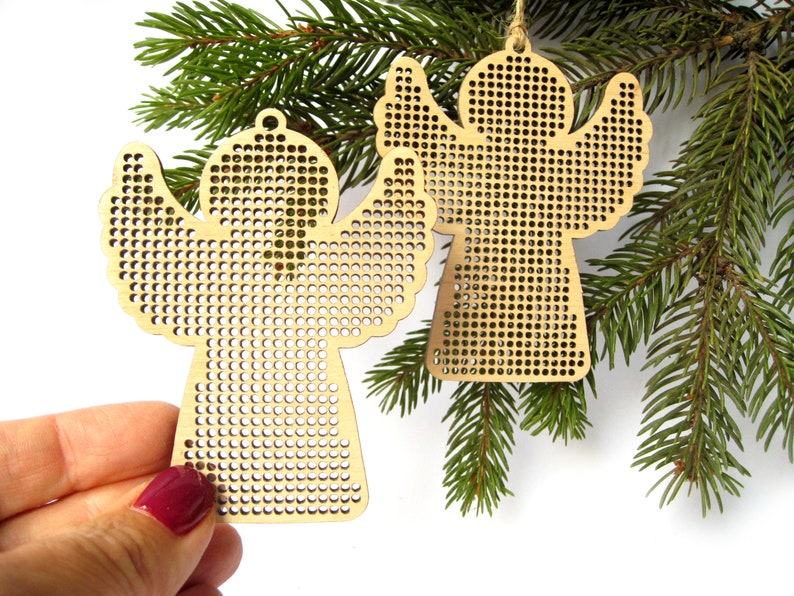 May include: Two wooden angel ornaments with perforated designs, hanging from twine, displayed on a green pine branch. The angels have outstretched wings and are made of light-colored wood. One is held by a hand with a maroon nail polish.