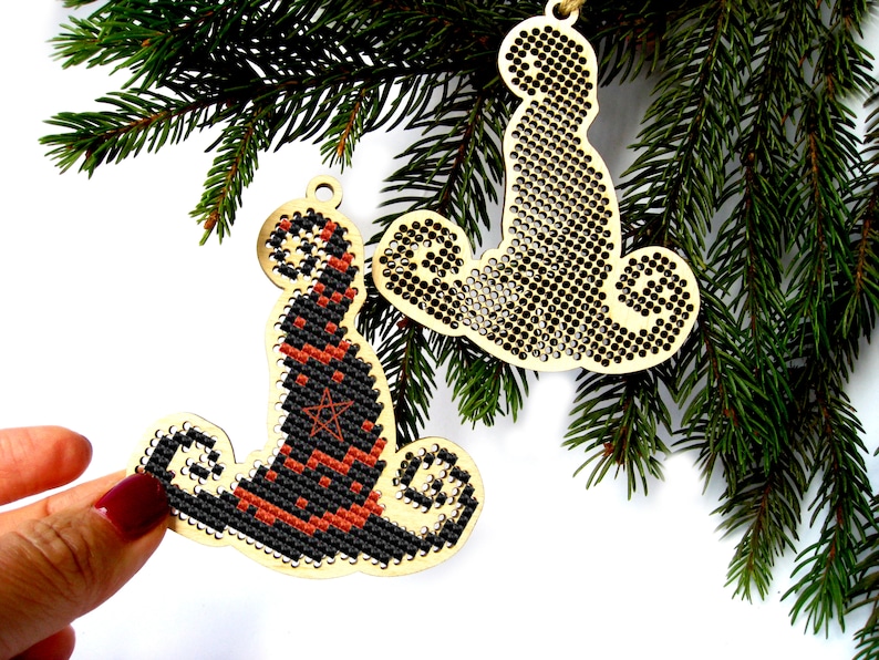 May include: Two wooden Christmas ornaments, one shaped like a witch's hat and the other like a cat. Both ornaments are decorated with black and orange cross-stitch designs.