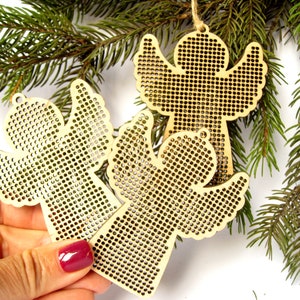 May include: Three wooden angel ornaments with a perforated design, hanging from twine. The angels are a light tan color and are displayed with evergreen branches in the background. The ornaments are suitable for Christmas or holiday decor.