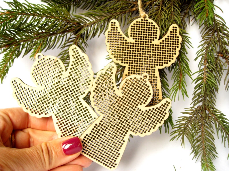 May include: Three wooden angel ornaments with a perforated design, hanging from twine. The angels are a light tan color and are displayed with evergreen branches in the background. The ornaments are suitable for Christmas or holiday decor.