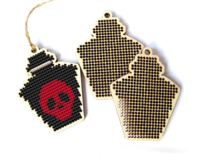 May include: Two wooden ornaments shaped like bottles with a grid pattern for cross-stitch. One ornament is decorated with a black and red skull.