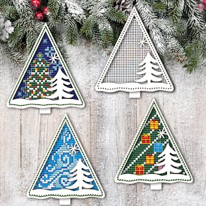 May include: Four Christmas tree ornaments with different cross-stitch designs. The trees are triangular with a white cutout tree and base. Designs include a Christmas tree, snowflakes, and wrapped gifts. The background is a wooden surface with evergreen branches.