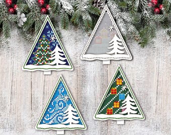 May include: Four Christmas tree ornaments with different cross-stitch designs. The trees are triangular with a white cutout tree and base. Designs include a Christmas tree, snowflakes, and wrapped gifts. The background is a wooden surface with evergreen branches.