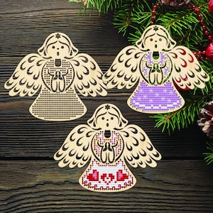 Peut inclure: Trois ornements en bois en forme d'ange avec des motifs d&eacute;coup&eacute;s complexes. L'un est uni, un autre pr&eacute;sente un motif de point de croix violet, et le troisi&egrave;me a un motif de point de croix rouge et blanc. Les anges sont pr&eacute;sent&eacute;s sur une surface en bois fonc&eacute; avec des d&eacute;corations de No&euml;l.
