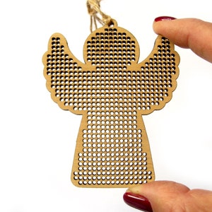 May include: A wooden angel ornament with a perforated design. The angel has outstretched wings and a small loop for hanging. The ornament is held by a hand with red nail polish against a white background. The angel is made of light brown wood.