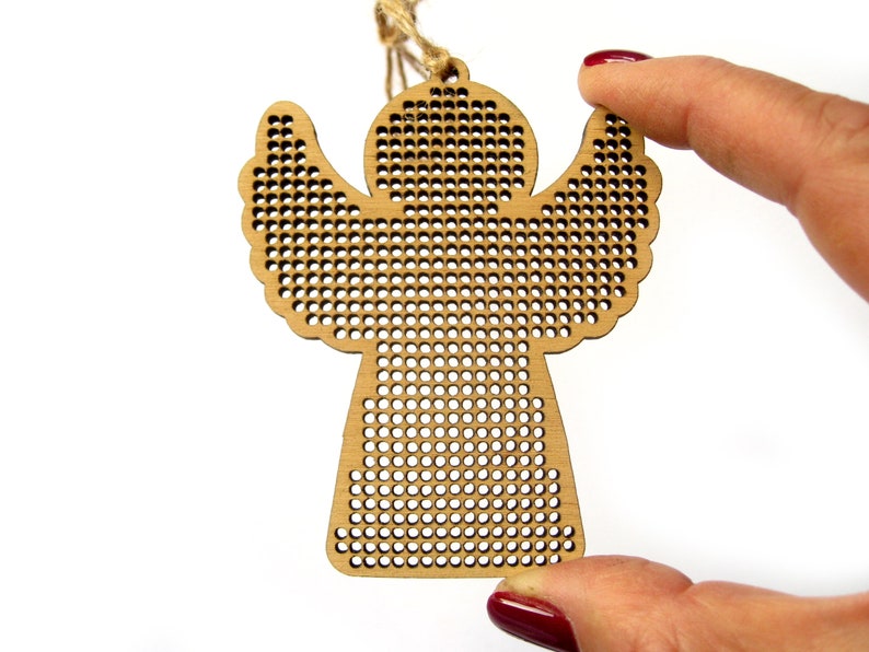 May include: A wooden angel ornament with a perforated design. The angel has outstretched wings and a small loop for hanging. The ornament is held by a hand with red nail polish against a white background. The angel is made of light brown wood.