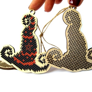 May include: Two wooden Halloween ornaments, one with a black and orange witch's hat and the other with a black cat design. Both ornaments are decorated with black and white cross-stitch patterns.