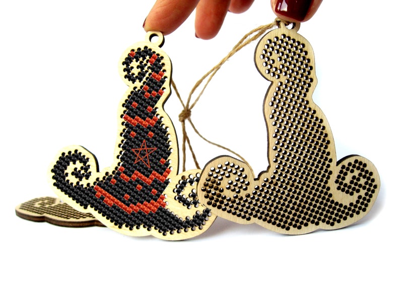May include: Two wooden Halloween ornaments, one with a black and orange witch's hat and the other with a black cat design. Both ornaments are decorated with black and white cross-stitch patterns.