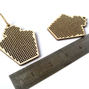 May include: Two unfinished wooden ornaments shaped like houses with a grid pattern for cross-stitching. The ornaments are approximately 2 inches wide and are shown next to a ruler.