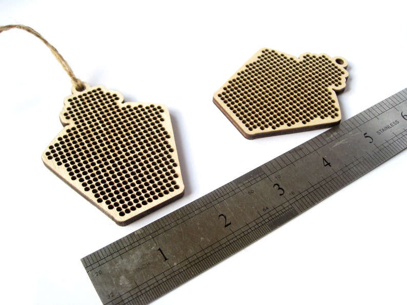 May include: Two unfinished wooden ornaments shaped like houses with a grid pattern for cross-stitching. The ornaments are approximately 2 inches wide and are shown next to a ruler.