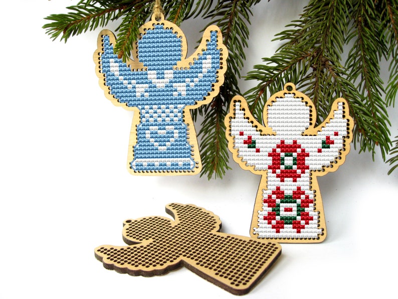 May include: Wooden angel ornaments with cross-stitch designs in blue and red, with a blank wooden angel ornament. The angels are hanging from a green pine branch, perfect for Christmas decorations.