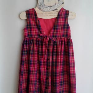 Puede incluir: Un vestido sin mangas de cuadros rojos, negros y amarillos, con cierre de botones en la parte delantera y un lazo en la espalda. El vestido est&aacute; colgado en una percha de madera con una ilustraci&oacute;n de rat&oacute;n.