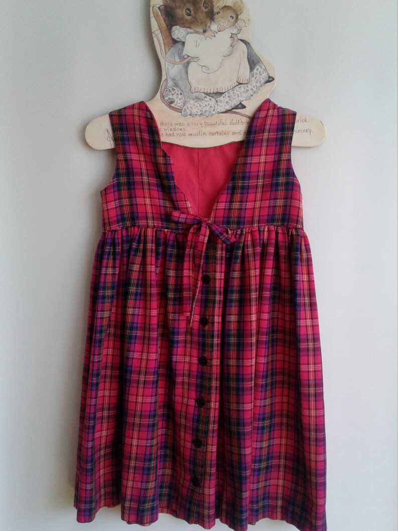 Puede incluir: Un vestido sin mangas de cuadros rojos, negros y amarillos, con cierre de botones en la parte delantera y un lazo en la espalda. El vestido est&aacute; colgado en una percha de madera con una ilustraci&oacute;n de rat&oacute;n.