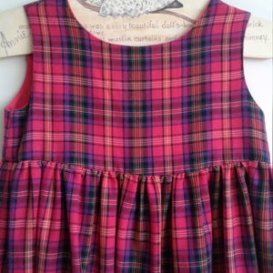 Puede incluir: Un vestido sin mangas de cuadros rojos y negros con falda fruncida. El vestido est&aacute; colgado en una percha de madera.
