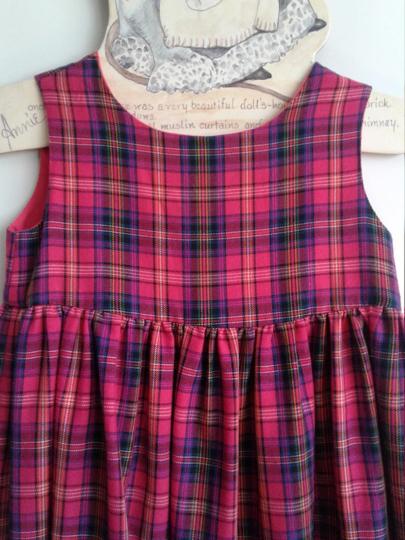 Puede incluir: Un vestido sin mangas de cuadros rojos y negros con falda fruncida. El vestido est&aacute; colgado en una percha de madera.