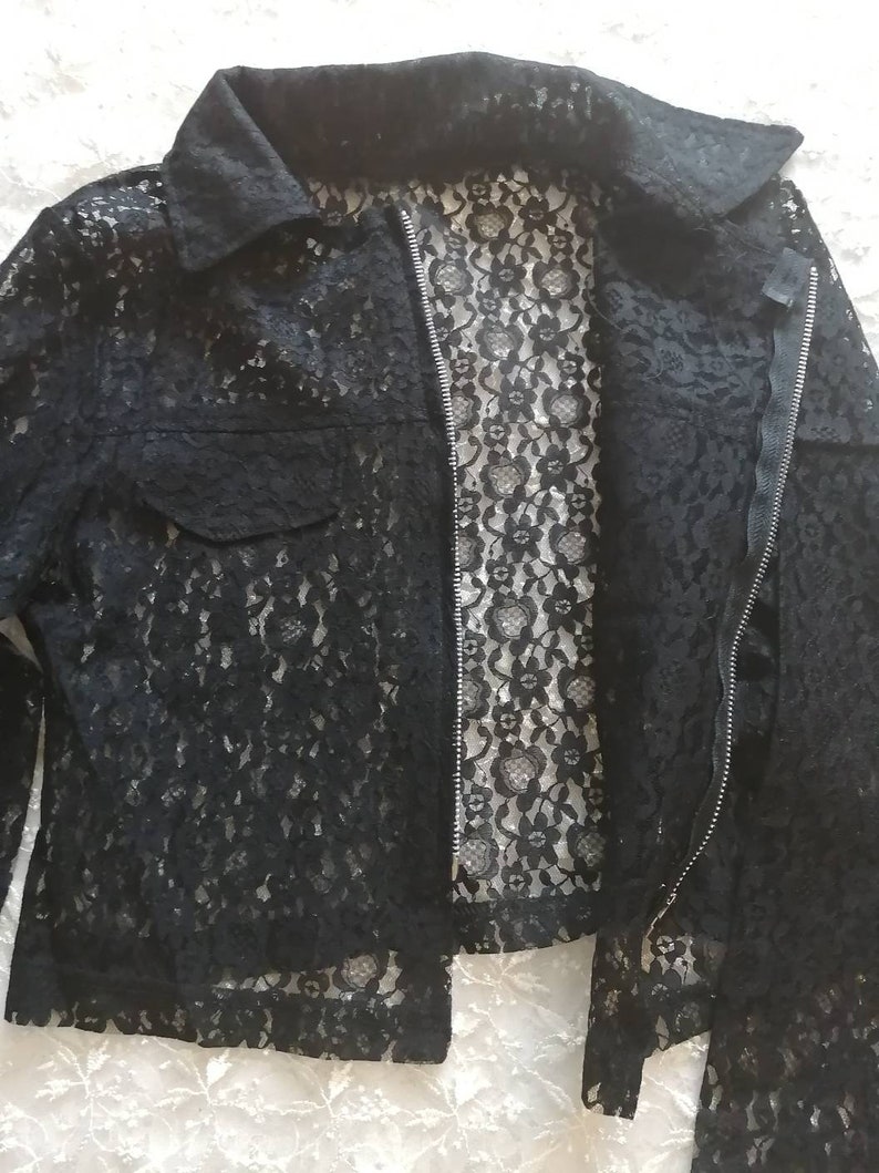 May include: Black lace jacket with a collared neckline, a front zipper, and a single pocket. The jacket's intricate floral lace pattern is visible throughout, creating a delicate and textured appearance. The jacket is a stylish outerwear option.