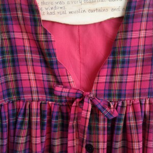 Puede incluir: Un vestido sin mangas de cuadros rojos y negros con un lazo en la cintura y un cierre de bot&oacute;n. El vestido tiene un escote en V y una falda fruncida.