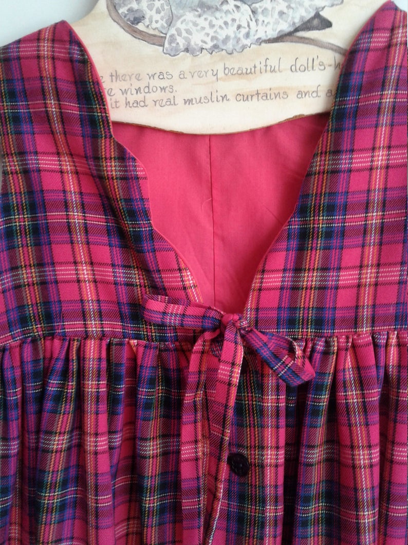 Puede incluir: Un vestido sin mangas de cuadros rojos y negros con un lazo en la cintura y un cierre de bot&oacute;n. El vestido tiene un escote en V y una falda fruncida.