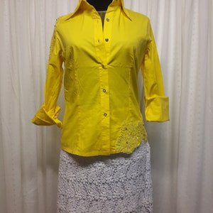 Peut inclure: Une chemise boutonnée jaune avec une jupe en dentelle blanche. La chemise a un détail décoratif sur le côté gauche et des boutons argentés.