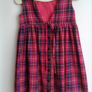 Puede incluir: Un vestido sin mangas de cuadros rojos y negros, con botones en la parte delantera y un lazo en la cintura.