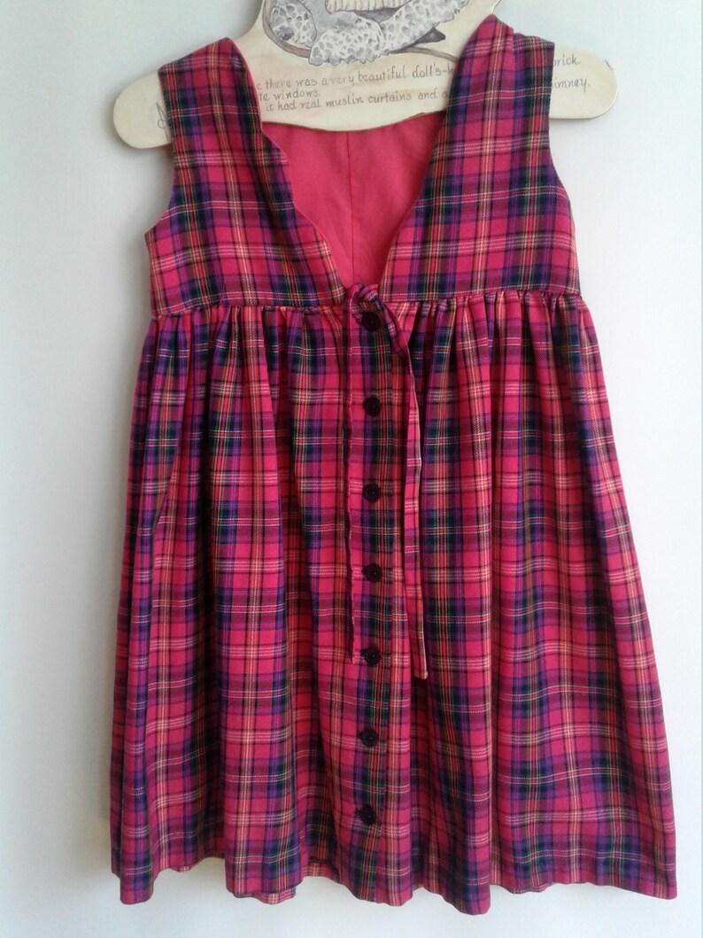 Puede incluir: Un vestido sin mangas de cuadros rojos y negros, con botones en la parte delantera y un lazo en la cintura.
