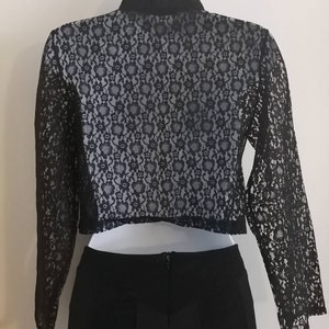 May include: Black lace bolero jacket with long sleeves, paired with a black skirt. The bolero features a floral lace pattern over a light-colored lining. The jacket has a high collar and is hanging on a white hanger.