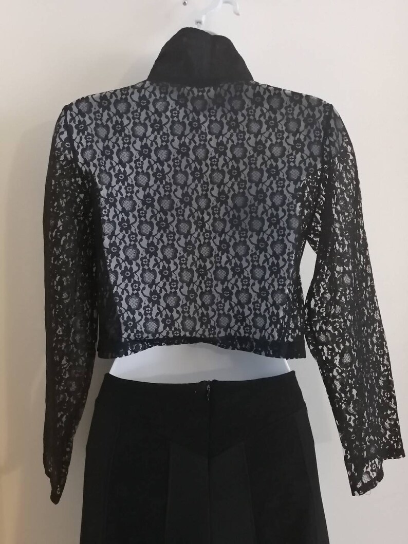May include: Black lace bolero jacket with long sleeves, paired with a black skirt. The bolero features a floral lace pattern over a light-colored lining. The jacket has a high collar and is hanging on a white hanger.