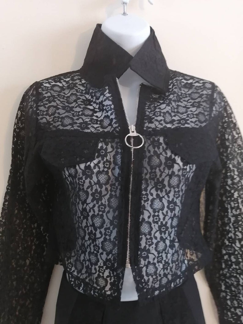 May include: Black lace jacket with a collared neckline and a silver zipper. The jacket features a floral lace pattern and two small pockets. The jacket is displayed on a white mannequin.
