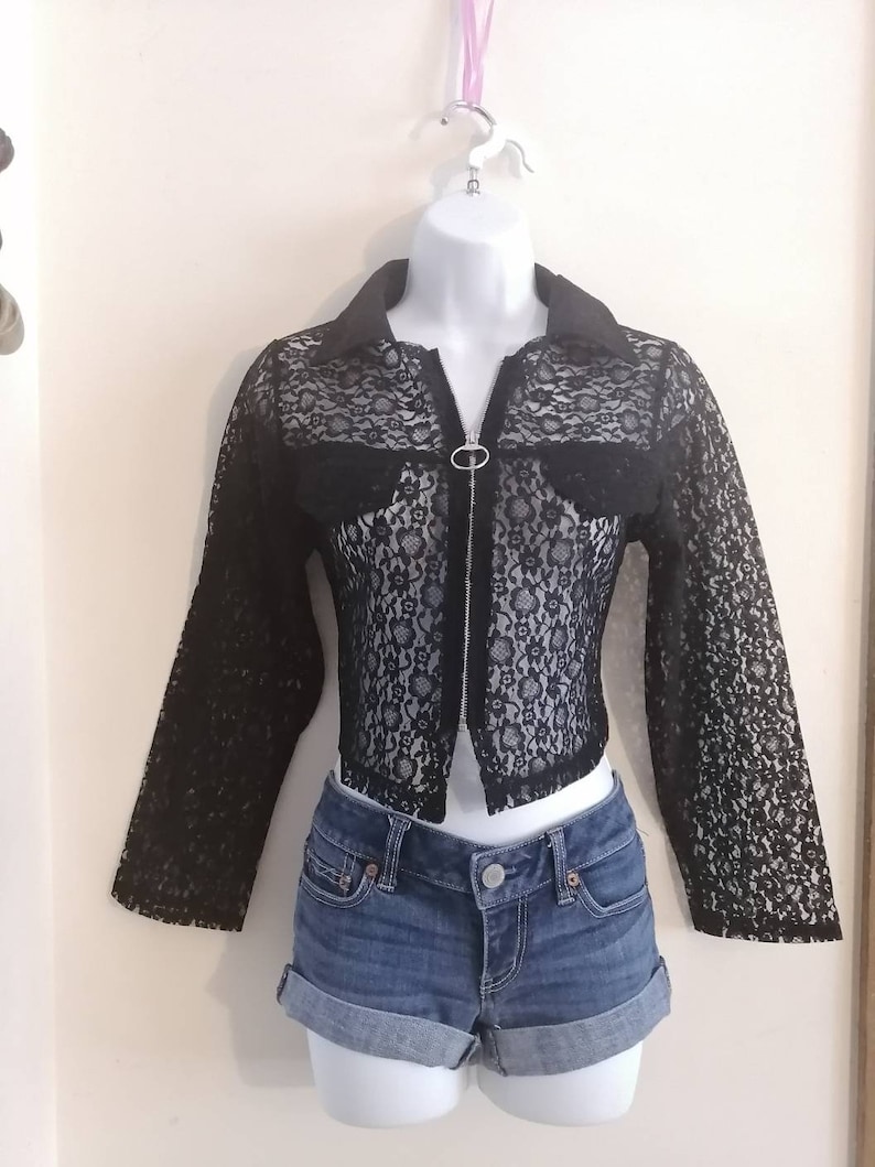 May include: A black lace jacket with a collar and long sleeves, paired with denim shorts. The jacket has a zipper closure and is displayed on a mannequin. The shorts are cuffed at the hem.