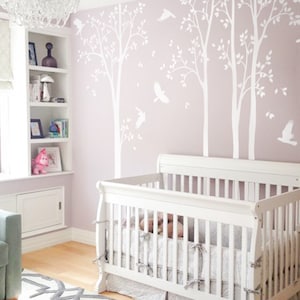 May include: A white cot with a grey and white patterned cot skirt and bedding. The nursery has a light purple wall with white tree decals and flying birds. A white chandelier hangs from the ceiling.