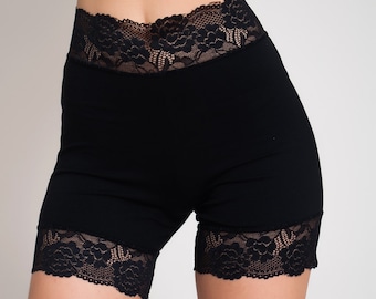 May include: Black high-waisted shorts featuring a wide floral lace waistband and matching floral lace trim along the bottom of each leg opening. The garment is constructed from a solid black fabric with a smooth texture and scalloped lace edges.