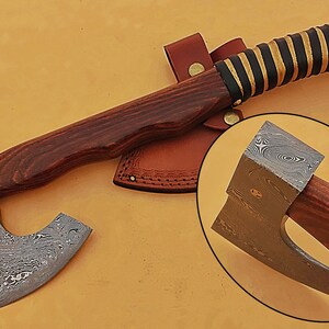 May include: A hand-forged Damascus steel axe with a wooden handle and a leather sheath. The axe has a unique pattern on the blade and a brass rivet.