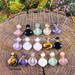 Può includere: Una collezione di collane con ciondoli di pietre preziose in vari colori, tra cui rosa, nero, verde e viola. Ogni ciondolo ha la forma di una piccola bottiglia sfaccettata con una catena d'argento o d'oro. Le collane sono esposte su una superficie di legno strutturata. Il testo "Forbeads" e un indirizzo web sono visibili.