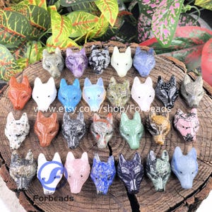 Könnte beinhalten: Eine Sammlung von geschnitzten Stein-Fuchs-Kopf-Figuren in verschiedenen Farben, darunter Rot, Weiß, Blau und Grau. Die Figuren werden auf einer Holzoberfläche mit grünem Laub im Hintergrund präsentiert. Der Text "Forbeads" ist sichtbar.