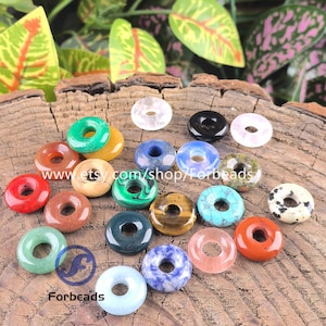 May include: A collection of gemstone donut beads in a variety of colours, including red, green, blue, and brown. The beads are arranged on a rustic wooden surface. The Etsy shop name "Forbeads" is visible.