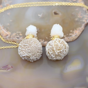 Może przedstawiać: Dwa złote łańcuszki z białym kryształem na górze i brązowym, kryształowym wisiorem w kształcie kuli.