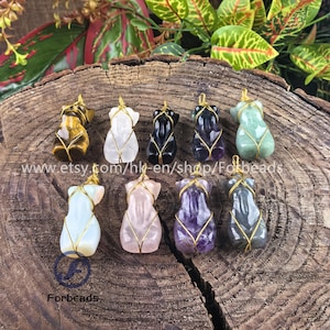May include: Ten different coloured gemstone pendants, each wrapped in gold wire, are displayed on a wooden surface. The gemstones include tiger's eye, clear quartz, black obsidian, amethyst, green aventurine, and more.