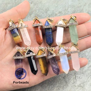 May include: A collection of twelve hexagonal crystal point pendants in various colors including blue, clear, red, black, yellow, purple, and pink. Each pendant features a gold-tone metal cap encrusted with small clear rhinestones and a bail.