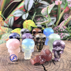 May include: A selection of small, carved crystal figurines in shades of pink, blue, and purple. The figurines are in the shape of a stylized alien or owl. The background features green and pink foliage, and the Etsy shop name is visible.