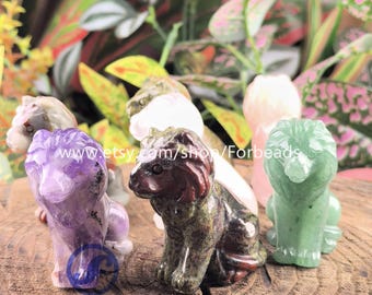May include: A selection of carved gemstone lion figurines in shades of purple, green, and pink. The Etsy shop name "Forbeads" is visible. The figurines are presented on a wooden surface. The background features foliage.