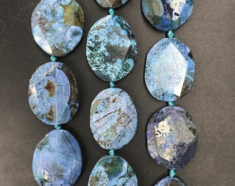 May include: A strand of blue agate gemstone beads with a faceted oval shape. The beads have a mottled blue and gray pattern with white and brown accents.
