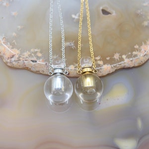 Może przedstawiać: Dwa srebrne i złote naszyjniki z przezroczystymi szklanymi wisiorkami w kształcie butelek. Butelki mają srebrne i złote korki.