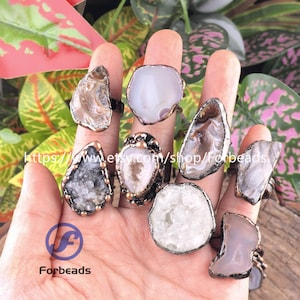 May include: A hand shows several gemstone rings with unique, irregular shapes. The rings feature various colours, including white, grey, and brown, set in bronze-coloured metal. The rings are displayed against a backdrop of green and pink foliage. The Etsy shop name "Forbeads" is visible.