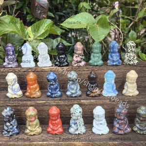 Puede incluir: Una colección de 20 pequeñas estatuillas de Buda talladas a mano en piedra, en varios colores y estilos. Las estatuillas están dispuestas en tres filas en un estante de madera.