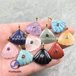Pode incluir: Uma coleção de pingentes de pedras preciosas em várias cores, incluindo azul, roxo, rosa, verde e laranja. Cada pingente tem uma argola dourada e um pequeno detalhe de pedra preciosa redonda. Os pingentes são em forma de leque e parecem esculpidos.