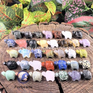May include: A collection of carved stone turtle figurines in various colors, including green, purple, and black. The turtles are displayed on a wooden surface. The Etsy shop name "Forbeads" is visible.