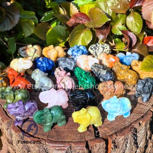 Könnte beinhalten: Eine Sammlung kleiner, geschnitzter Elefantenfiguren aus Stein in verschiedenen Farben, darunter Grün, Rosa, Gelb und Blau. Die Elefanten sind auf einer Holzoberfläche mit grünem und rotem Laub im Hintergrund ausgestellt. Der Etsy-Shop-Name "Fonbeads" ist sichtbar.
