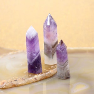 May include: Three amethyst crystal points of varying sizes, with purple and white banding, are displayed on a light brown agate slice.